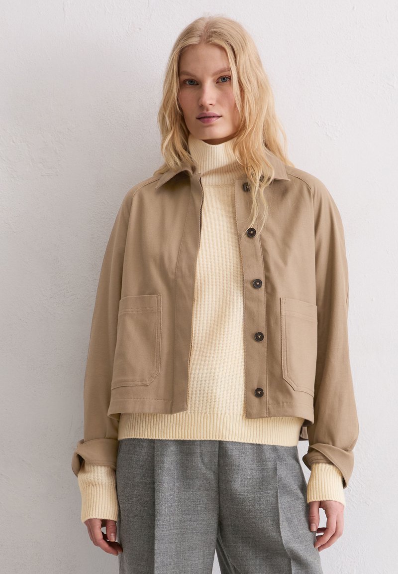 Krótkia kurtka w kolorze beżowym z dwoma przednimi kieszeniami, brązowymi guzikami, noszona na kremowym golfie z prążkowanej dzianiny oraz szare spodnie w kratę. Miękka faktura tkaniny.