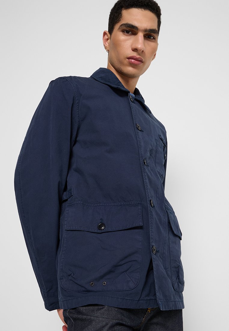 Giacca blu navy con chiusura a bottoni, due tasche frontali e colletto. Realizzata in tessuto leggero, presenta una vestibilità comoda e un design semplice.