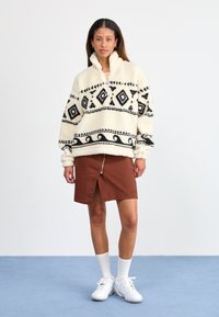 Jonge vrouw draagt een crèmekleurige fleece jas met een patroon, een bruine rokje met rits aan de voorkant, witte sokken en witte sneakers, staande op een blauwe vloer.