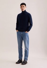 Pull à col roulé bleu marine, coupe ajustée, porté avec un jean bleu clair et des chaussures noires. Texture lisse, design simple et contraste minimal des couleurs.