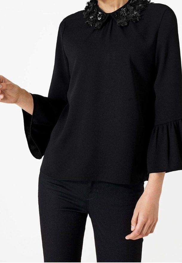 Femme portant un chemisier noir à manches longues avec des manches cloche à volants et un col floral orné, associé à un pantalon noir ajusté.