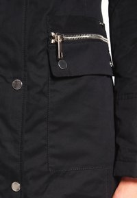 Svart, isolerad jacka med matt yta. Har en ficka på bröstet med dragkedja, silverfärgad hårdvara och tryckknappsdetaljer.