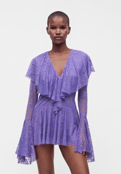 Femme portant une courte robe en dentelle violette avec de longues manches évasées et un col volanté en V profond, debout devant un fond blanc uni.