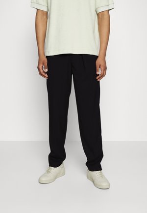 Persona che indossa pantaloni neri a vestibilità ampia, sneaker color avorio e una camicia verde chiaro a maniche corte, in piedi davanti a uno sfondo semplice.