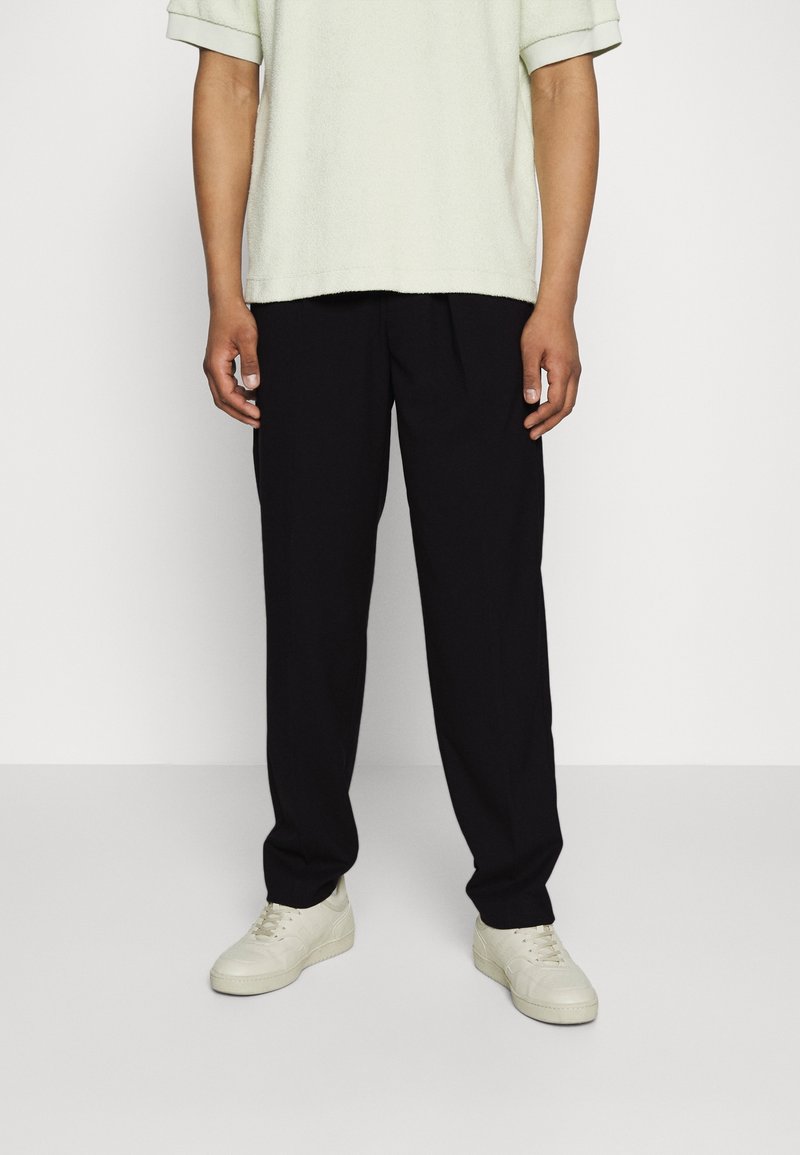 Personne portant un pantalon noir ample, des baskets écrues et une chemise à manches courtes vert pâle, debout devant un fond uni.