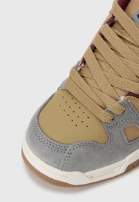 Chaussure de sport avec une tige en suède gris, un bout renforcé synthétique beige, des lacets marron et des perforations pour la ventilation. Semelle extérieure en caoutchouc visible.