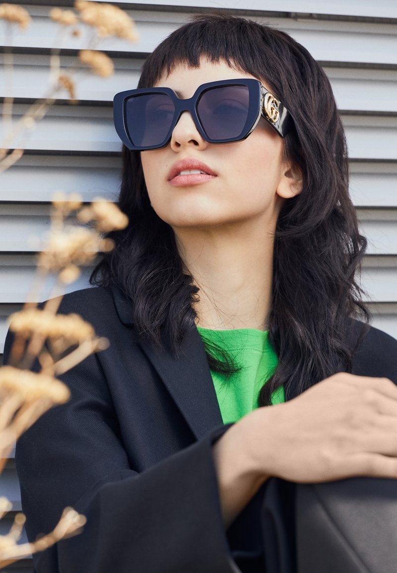 Ochelari de soare supra-dimensionați, de culoare neagră, cu un cadru gros și accente aurii, purtați cu un sacou negru și o cămașă verde aprins, pe un fundal orizontal.