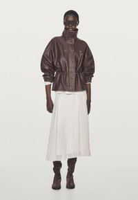 Brun läderjacka med hög krage, åtdragen midja; vit, flödande kjol; knähöga bruna stövlar. Modellen bär solglasögon.