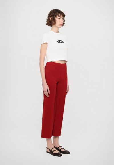 T-shirt blanc coupé avec logo noir, associé à un pantalon rouge taille haute et à large jambe. Chaussures à talons ouverts noires avec des accents décoratifs en métal.