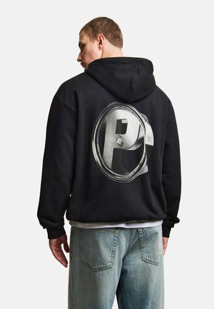 Mężczyzna w czarnej bluzie z kapturem z dużym metalicznym, trójwymiarowym wzorem litery „P” na plecach, w zestawieniu z jasnoniebieskimi dżinsami, stojący tyłem do kamery.