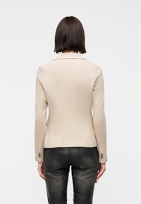 Femme aux cheveux raides et foncés portant un blazer beige cintré et un pantalon en cuir noir, vue de dos sur un fond uni.