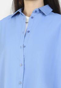 Camisa azul claro con un cuello clásico, que cuenta con un frente abotonado y botones azules; textura suave, diseño casual.
