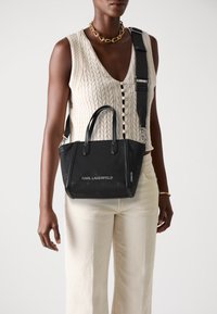 Black handbag with dual handles, adjustable strap, smooth material, embossed logo, and a structured shape. Worn by a model in casual attire.