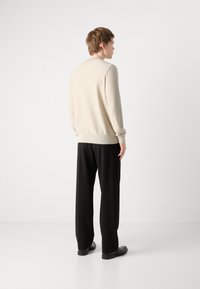 Beige neulepusero, jossa on ribbireunat hihoissa ja helmassa, yhdistetty mustiin leveälahkeisiin housuihin ja mustiin kenkiin, nähtynä takaa.