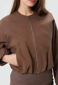 Bruine korte bomberjack met een ritssluiting aan de voorkant, elastische manchetten en een aansluitende zoom. Gemaakt van gladde stof met een matte textuur.