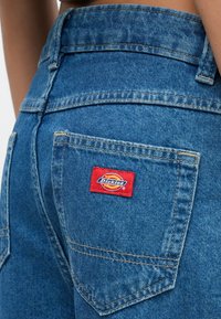 Κοντινή λήψη πίσω τσέπης μπλε τζιν με κόκκινο ορθογώνιο λογότυπο και ορατές ραφές στις άκρες της μέσης και της τσέπης.