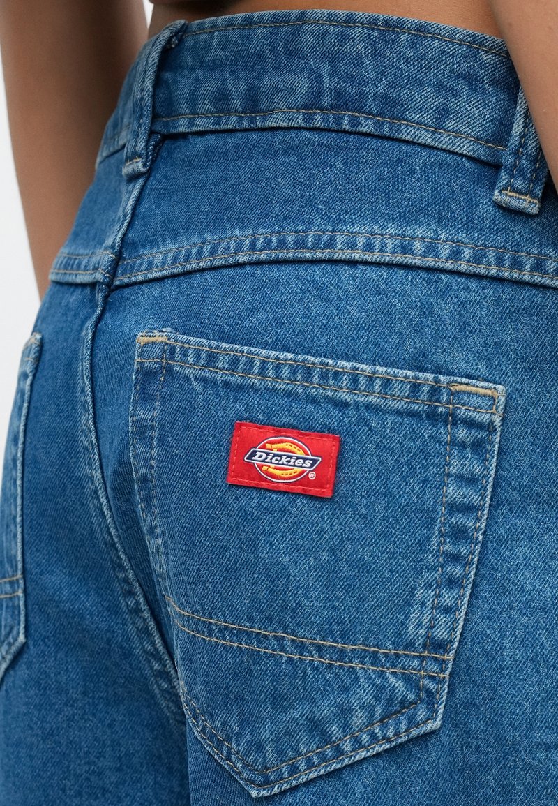 Close-up van de achterzak van een blauwe spijkerbroek met een rood rechthoekig logoplaatje en zichtbare stiksels langs de taille- en zakranden.