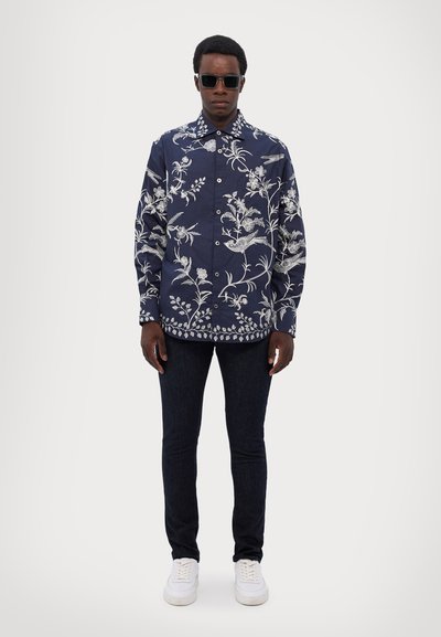 Camisa de botones floral en azul marino con bordado botánico blanco, combinada con jeans oscuros y zapatillas blancas. El modelo usa gafas de sol.