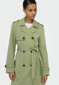 Femme portant un trench-coat à double boutonnage vert clair avec ceinture à la taille, poignets boutonnés et épaulettes, regardant légèrement vers le bas.