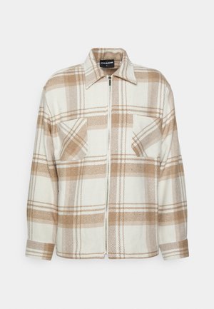 Mężczyzna w beżowo-białej kratowanej kurtce zapinanej na zamek, czarnej koszulce, ciemnoszarych dżinsach, białych i czarnych sportowych butach oraz srebrnych naszyjnikach łańcuszkach.