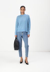 Hellblau gestrickter Pullover mit einem strukturierten Muster, kombiniert mit hochtaillierten Jeans aus Denim und schwarzen Lackabsätzen. Schwarze Handtasche wird in der Hand gehalten.