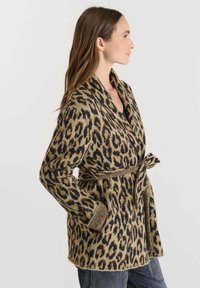 Cárdigan de punto con estampado de leopardo, mangas largas, cuello chal y cintura anudada. Base texturizada en beige con manchas negras. Material grueso y acogedor.