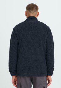 Giacca in fleece di colore blu navy con una superficie testurizzata e morbida; presenta un colletto alto e maniche lunghe. L'orlo è leggermente curvato nella parte inferiore.