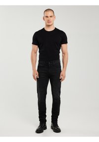 Bărbat stând cu fața spre înainte, purtând un tricou negru, blugi negri și bocanci negri cu șireturi, pe un fundal simplu deschis la culoare.