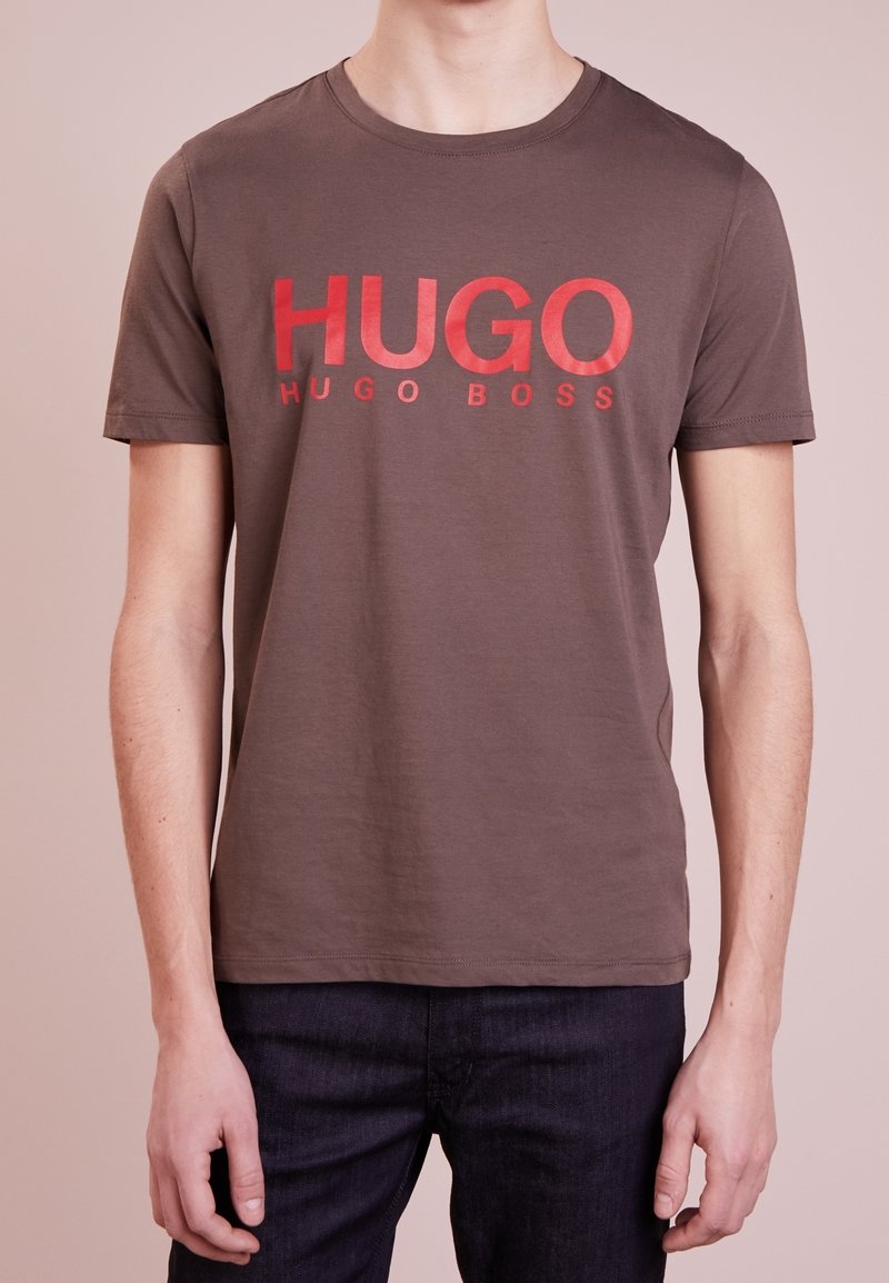 Camiseta de algodón marrón con cuello redondo, que presenta el texto en rojo brillante "HUGO" en el pecho y "HUGO BOSS" debajo en un tamaño de fuente más pequeño.