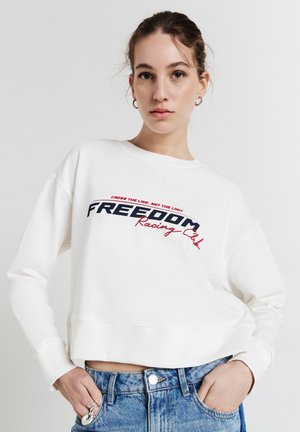 Giovane donna che indossa una felpa bianca con la scritta "FREEDOM Racing Club" e jeans blu, in posa con le mani nelle tasche su uno sfondo semplice.