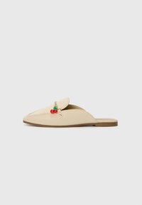 Zapatos slip-on beige con parte superior de tejido, suela plana y un charm decorativo de cereza en rojo y verde en la parte delantera. Textura suave, diseño minimalista.