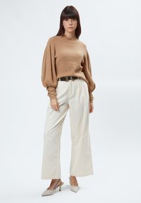Beiger gerippter Pullover mit hohem Kragen, voluminösen Puffärmeln; helle weit geschnittene Hose; helle spitz zulaufende High Heels mit strukturiertem Finish.