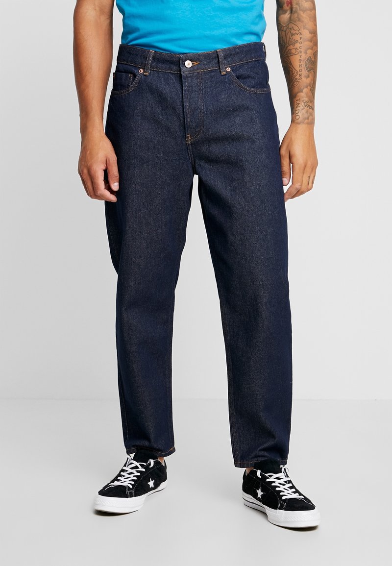 Homme portant un jean droit bleu foncé, des baskets noires avec des lacets blancs et une chemise bleu vif, sur un fond uni.