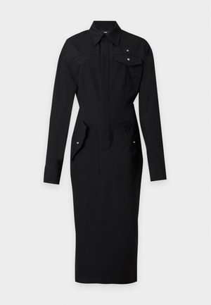 Femme aux longs cheveux noirs portant une robe-chemise noire à manches longues avec plusieurs poches et des chaussures noires à talons hauts, debout sur un fond blanc.