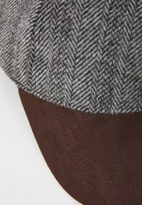 Căciulă cu model houndstooth, având un vârf din țesătură gri și neagră și o vizieră solidă din țesătură maro, prezentând detalii de cusătură vizibile.