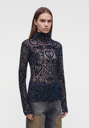 Femme portant un haut col roulé noir transparent à manches longues avec des motifs complexes de cœurs et d’étoiles, associé à un pantalon bicolore avec de grandes poches à l’avant.