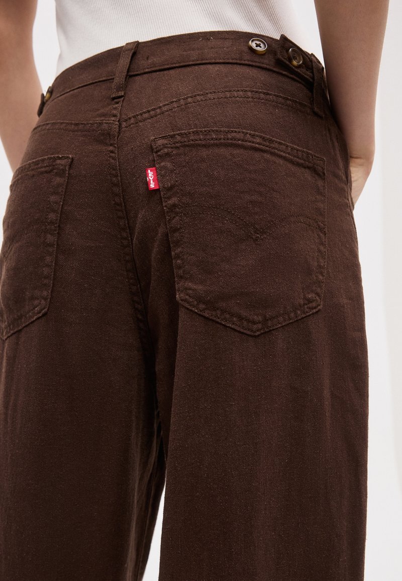 Person wearing high-waisted dark brown Levi's jeans, showing back pockets with red Levi's tag and button details at waistband.