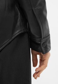 Giacca di pelle nera con un design elegante, texture morbida e chiusura a bottone sulla manica, abbinata a un tessuto scuro sotto.