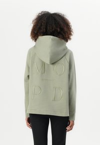 Person med krøllet hår iført en afdæmpet grøn hættetrøje med store præget bogstaver M, O, P, D og en lille tekst "MARCO POLO DENIM" på ryggen.