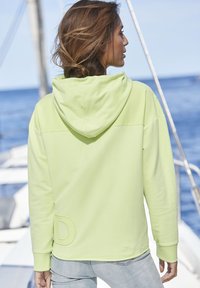 Lime green hoodie with a soft texture, kangaroo pocket, and embroidered ‘D’ detail on the left side, worn with light blue jeans.