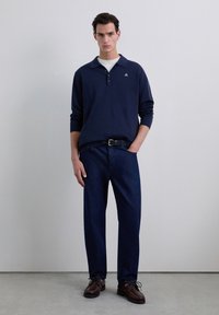 Polo marin cu mânecă lungă, cu guler și plăcuță cu butoane, asortat cu blugi drepți de culoare albastru închis și pantofi maro cu siret. Design simplu.
