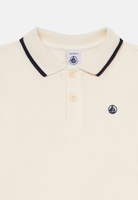 Kreminės spalvos kūdikių polo marškiniai su tamsiai mėlynu apvadu ant apykaklės, dviem mygtukais ir mažu tamsiai mėlynu burlaivių logotipu ant krūtinės, 12 mėnesių dydžio.