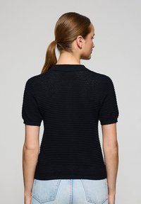Polo nero in maglia con maniche corte, caratterizzato da un motivo testurizzato e polsini a costina. La vista posteriore mostra una silhouette aderente e dettagli del colletto.