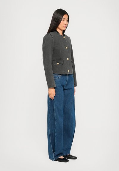 Jongeman staat zijwaarts met een donkergrijze knoopjas, wijde blauwe jeans en zwarte platte schoenen op een witte achtergrond.