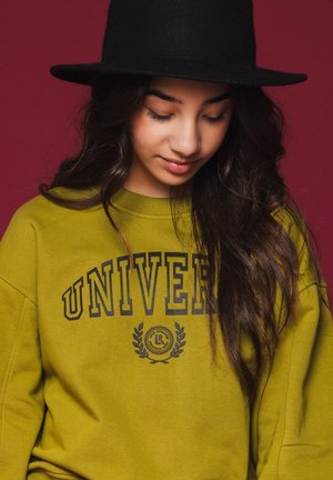 Olijfgroene sweatshirt met "UNIVER" in zwart gedrukt, met een ronde halslijn en een losse pasvorm. Zwarte vilten hoed gedragen op lang, golvend haar.