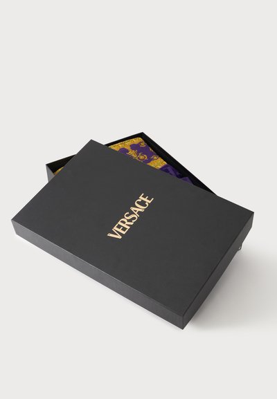Boîte noire Versace avec logo doré partiellement ouverte, révélant un tissu violet et doré à motifs à l'intérieur.