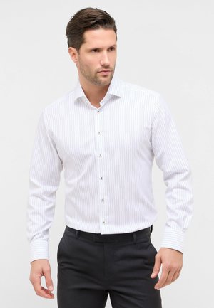 Uomo con indosso una camicia elegante bianca slim fit a righe verticali sottili e pantaloni eleganti neri, in piedi davanti a uno sfondo semplice.