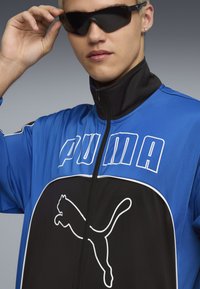 Zila un melna sporta jaka ar augstu apkakli, priekšējo rāvējslēdzi, izcelto "PUMA" logo un baltu kaķa siluetu uz priekšpuses.
