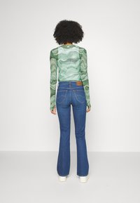 Top verde de manga larga con un diseño de espiral y textura transparente, combinado con jeans acampanados azules que cuentan con bolsillos traseros y un parche de cuero con marca.