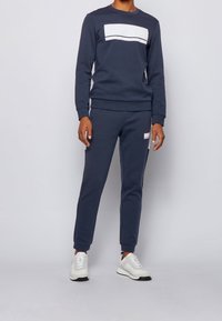 Granatowy komplet dresowy z bluzą z okrągłym dekoltem i białymi paskami oraz dopasowanymi zwężanymi spodniami. Materiał wydaje się miękki i o fakturze.